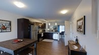 This two-storey space showcases a well-designed open-concept main floor where the kitchen, dining, and living areas flow seamlessly into one another. The kitchen is a standout feature, equipped with dark espresso cabinetry that extends to the upper walls, stainless steel appliances including a French door refrigerator, microwave, and range, complemented by a dishwasher integrated into the lower cabinetry. A peninsula with a dark countertop serves as a natural divider between the kitchen and the adjoining living area, providing additional workspace and casual seating potential. Pendant lighting fixtures hang above the peninsula, adding focused illumination and a decorative touch. The living area beyond benefits from natural light filtering through a window dressed with blue curtains, creating a bright and inviting atmosphere. Warm hardwood-style flooring runs throughout the main level, adding cohesion and a sense of warmth to the space. A patterned area rug in the kitchen zone adds texture underfoot. The layout is practical and open, allowing for easy movement between functional zones, while flush-mount ceiling lighting ensures the entire area remains well-lit throughout the day.
