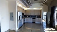This single-floor space showcases a compelling blend of industrial and contemporary design elements that come together to create a stylish, urban atmosphere. The open-plan kitchen serves as a central feature, outfitted with a full suite of stainless steel appliances, including a French door refrigerator with built-in water dispenser, a range with oven, a built-in microwave, and a dishwasher. Light wood-toned upper cabinetry paired with crisp white lower cabinets creates a striking two-tone aesthetic, while dark countertops and a dark mosaic tile backsplash add depth and contrast. Ambient LED lighting installed beneath a tiered ceiling soffit bathes the kitchen in a warm, layered glow, enhancing the modern character of the space. Overhead, exposed industrial ductwork runs across the high ceiling, reinforcing the loft-inspired aesthetic. The polished concrete flooring extends throughout, offering a sleek, low-maintenance surface that reflects light and contributes to the open, airy feel of the space. Large windows allow natural light to pour in, connecting the interior to the outside environment. The overall design strikes a balance between raw industrial elements and refined modern finishes, making for a distinctive and functional living space.