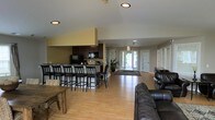 This single-story space showcases an open-concept layout that seamlessly integrates a kitchenette, dining area, and lounge into one cohesive environment. The kitchenette is equipped with dark espresso cabinetry, countertop appliances including a microwave and coffee maker, and is anchored by a long bar-height counter lined with black bar stools — providing ample casual seating for group gatherings. The vaulted ceiling in the kitchen area adds a sense of height and openness to that portion of the room.

The lounge area features dark leather sofas arranged for comfortable seating, complemented by a rustic wooden coffee table. Light hardwood flooring runs throughout the entire space, contributing to a warm and inviting atmosphere. A large area rug defines the transition between the seating and entry zones.

Natural light flows generously through a set of glass-paneled double doors at the far end of the room, as well as through adjacent sidelights and windows. The entry area features a welcome mat and a small console table, lending a welcoming feel. Recessed lighting throughout ensures the space is well-lit at any time of day. Overall, the layout is well-suited for communal or multi-purpose use.