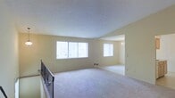 This single-floor space offers an open, airy layout with a seamless flow between the main living area and an adjoining dining or secondary room. The interior is finished with soft, wall-to-wall carpeting throughout, creating a warm and comfortable atmosphere underfoot. Neutral, light-toned walls reflect natural light generously, contributing to the overall brightness of the space.

The main living area is illuminated by a wide bank of windows dressed with vertical blinds, allowing for flexible light control while maintaining a clean, classic look. A textured ceiling adds a subtle visual element to the expansive overhead space. A wrought iron railing detail is visible, hinting at a split-level or elevated entry design that adds architectural character.

The adjoining room, accessible through a wide open archway, features its own window with vertical blinds and is accented by an elegant chandelier overhead, lending a touch of sophistication. The open-plan design between these two spaces promotes a sense of generous square footage, making the interior feel connected and easy to navigate.

Overall, the space presents a clean, neutral palette that is move-in ready and well-suited for a variety of furnishing styles, with natural light and an open layout as its defining features.