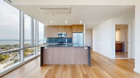Located at 50 Clinton Pl in New Rochelle, NY, this high-rise unit offers a refined, contemporary living experience across a single thoughtfully designed floor. The open-concept layout is anchored by a sleek, modern kitchen outfitted with wood-grain cabinetry, dark stone countertops, stainless steel appliances including a French door refrigerator and built-in microwave, and a central island with pendant lighting overhead. Light-toned hardwood flooring runs throughout, lending warmth and continuity to every room. A defining architectural feature of this unit is its extensive floor-to-ceiling glass curtain wall, which frames sweeping, elevated views of the New Rochelle cityscape and Long Island Sound beyond. Natural light pours into every room, creating a bright, airy atmosphere throughout the day. A prominent curved column in the living area adds a striking architectural accent against the panoramic backdrop. The bedroom spaces benefit directly from the expansive glazing, each offering unobstructed water and city views. The bathroom is finished with large-format porcelain tile flooring, a wood-tone vanity with a stone countertop, gray marble-style wall tile, and a frameless glass-enclosed walk-in shower with brushed nickel fixtures — delivering a clean, spa-like aesthetic.