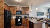 This single-floor residence combines modern functionality with warm, inviting design. The kitchen is a standout feature, showcasing rich knotty alder cabinetry, granite countertops with stone-tile backsplash, and a full suite of black appliances including a range, microwave, refrigerator, and dishwasher. A peninsula with seating and pendant lighting overhead creates a natural gathering space that opens seamlessly to the dining and living areas. Dark hardwood-style flooring runs throughout the main living spaces, adding warmth and continuity.

The living room is bright and comfortable, benefiting from natural light through large windows with access to an exterior door. A ceiling fan adds a practical touch, while the open layout allows for versatile furniture arrangements.

Both bedrooms are carpeted and well-lit, each with large windows that invite natural light and views of the surrounding greenery. Ceiling fans and ample wall space make them functional and comfortable retreats. The primary bedroom features stylish wall-mounted lighting flanking the bed area, lending a boutique hotel-like ambiance.

The bathroom continues the granite countertop theme, complemented by warm wood cabinetry and a well-organized vanity area. Overall, the space presents a cohesive, refined aesthetic with quality finishes throughout.