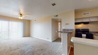 This single-floor 1-bedroom, 1-bathroom residence offers a practical and comfortable layout designed for everyday living. The living area is generously sized, featuring soft wall-to-wall carpeting, warm neutral tones, and a ceiling fan with integrated lighting. A large sliding glass door with vertical blinds floods the space with natural light and provides access to the outdoors. Adjacent to the living area, a half-wall peninsula with granite-style countertops delineates the kitchen, which features white cabinetry, dark countertops, a stainless steel sink, hardwood-style flooring, and black appliances including a dishwasher. The open sightlines between the kitchen and living space create a cohesive, connected feel throughout. The bedroom is a comfortable, carpeted retreat equipped with its own ceiling fan, a window with blinds for natural light, and clean, neutral walls. The bathroom is finished with classic white tile surrounding a full-size soaking tub and shower combination, complemented by basket-weave tile flooring and a mirrored medicine cabinet. Throughout the space, consistent warm beige tones, clean finishes, and thoughtful details contribute to a move-in-ready environment that balances functionality with comfort.