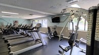 This single-level fitness center offers a well-organized workout space designed to accommodate a variety of exercise needs. The room features dark rubber flooring, which provides durability and traction underfoot, and is finished with a two-tone paint scheme of soft teal and gray that lends the space a clean, modern aesthetic. A textured ceiling with recessed lighting and fluorescent fixtures ensures consistent illumination throughout.

The cardio section lines one side of the room with multiple treadmills and an elliptical machine, while a stationary upright bike occupies the center of the space. A multi-function cable and strength training machine is prominently positioned toward the front, offering options for resistance-based workouts. A wall-mounted flat-screen TV is installed above eye level, allowing users to stay entertained during their sessions.

Natural light filters in through several windows along the walls, providing views of surrounding greenery and brightening the interior. Ventilation is supported by ceiling vents, contributing to a comfortable workout environment. Overall, the fitness center is efficiently laid out to maximize usable floor space while keeping equipment accessible.