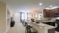 This single-floor residence showcases an open-concept layout that integrates the kitchen and living areas into one cohesive, functional space. The kitchen is outfitted with rich dark cabinetry, granite countertops, and a double sink, while an extended breakfast bar with bar-height seating provides a casual dining option and serves as a natural dividing element between the kitchen and living room. Recessed lighting and under-cabinet illumination ensure the kitchen remains well-lit throughout the day.

The living area features plush carpeting in a neutral tone, contributing to a warm and inviting ambiance. A ceiling fan with a light fixture enhances comfort and adds a classic design element overhead. A front entry door is flanked by a window dressed with layered curtains, allowing natural light to filter into the space. Neutral, beige-toned walls complement the furnishings and create a cohesive aesthetic throughout. A partial view into an adjacent room, likely a bedroom, is visible through an interior doorway, hinting at additional private spaces beyond the main living area.