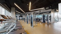 This expansive fitness center is designed to support a comprehensive range of training disciplines within a sleek, modern environment. The space is organized into distinct zones, each thoughtfully outfitted to meet diverse workout needs. The cardio area features a long row of treadmills and elliptical machines positioned along floor-to-ceiling glass walls, offering abundant natural light and open views of the surrounding cityscape. Adjacent to this, a dedicated strength training zone houses a robust selection of Star Trac cable machines, selectorized weight equipment, free-weight benches, and a full dumbbell rack. A functional training area features a multi-use suspension and battle-rope rig with hardwood flooring, alongside storage racks stocked with stability balls, medicine balls, and exercise mats. A separate studio space with hardwood floors and mirrored walls accommodates cycling bikes and group fitness sessions, with direct access to an outdoor terrace. Throughout the facility, a dark exposed ceiling with striking linear LED lighting creates a high-energy, industrial-modern ambiance. Durable rubberized flooring in the equipment zones and hardwood in the open training areas ensure both safety and aesthetic cohesion across the entire facility.
