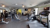 This fitness center offers a comprehensive workout space designed to support a variety of exercise routines. The main floor is finished with durable rubber flooring, providing a safe and practical surface for both strength training and cardio activities. A multi-station cable machine anchors the strength training area on one side of the room, complemented by additional weight equipment nearby. The cardio section features a row of stationary bikes and an elliptical trainer, positioned along the wall to allow ample space for movement. A wall-mounted flat-screen television is conveniently placed for entertainment during workouts. A ceiling fan helps maintain airflow throughout the space. One of the most distinctive design elements is the accent wall, adorned with motivational words and phrases — such as "commitment," "confidence," "results," and "energy" — rendered in bold green and white typography, adding visual energy to the room. A doorway leads to an adjacent area, visible in the background, which appears to offer additional equipment or amenities. Natural light filters in through a window, contributing to a bright and welcoming atmosphere overall.