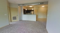 This single-floor space offers a contemporary open-concept layout that seamlessly connects the living area to the kitchen. The living space is finished with plush wall-to-wall carpeting, creating a warm and comfortable atmosphere, while flush-mount ceiling lighting ensures the area is well-lit throughout.

The kitchen is positioned at the heart of the unit, anchored by a peninsula with a light stone countertop that serves as both a workspace and a casual dining or entertaining surface. Dark cabinetry provides a striking contrast against the light walls, and the kitchen is equipped with stainless steel appliances, including a full-size refrigerator and built-in microwave. Pendant lighting hangs above the peninsula, adding a refined, modern touch.

A standout architectural feature is the exposed industrial-style ductwork that runs along the ceiling, lending the space a loft-inspired character. A wall-mounted HVAC vent near the entry ensures efficient climate control. The layout also reveals an additional room beyond the main living area, accessible through an open doorway, offering flexible use as a bedroom or office. The overall design balances industrial and modern aesthetics, making for a functional and visually engaging living environment.