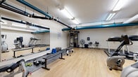 Located at 1396 Ogilvie Rd in Ottawa, Ontario, this fitness facility offers a fully equipped gym space designed to support a wide range of workout routines. The room features warm-toned hardwood-style flooring that contrasts with crisp white walls, creating a clean and functional environment. Overhead, exposed blue-painted pipes and bright fluorescent lighting run the length of the ceiling, giving the space an industrial character while ensuring excellent illumination throughout.

The gym is thoughtfully organized to maximize usability. A dedicated free weights area includes a collection of dumbbells in various sizes, a flat workout bench, and foam mat flooring for added comfort during floor exercises. A large wall-mounted mirror spans one side of the room, enhancing the sense of space and allowing users to monitor their form. 

On the cardio side, the facility is equipped with multiple treadmills, elliptical trainers, and a stationary cycle, providing diverse options for cardiovascular training. A multi-function cable machine anchors the strength training area, complemented by additional resistance equipment and a wall-mounted workout chart for reference.

A floor-standing fan and a wall-mounted device ensure comfort during intense sessions. The space is accessed through a solid door with a small interior window, and the overall layout allows for easy movement between equipment zones, making this gym a practical and well-appointed fitness destination.