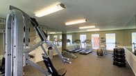 This fitness center is a dedicated single-floor workout space designed to accommodate a variety of exercise routines. The room is outfitted with multiple treadmills and an elliptical trainer, providing ample options for cardiovascular training. A weight bench and medicine ball round out the strength and cross-training equipment available to users. Full-length mirrors line one wall, enhancing the sense of space while allowing exercisers to monitor their form. Natural light filters in through a row of windows along the exterior wall, creating a bright and energizing environment. The floor is finished with durable rubber gym flooring, providing cushioning and grip suitable for high-impact activities. Overhead fluorescent lighting ensures the space remains well-lit throughout the day. The walls are painted in a neutral tone, contributing to a clean and focused atmosphere. The textured ceiling adds a practical finish to the utilitarian design of the room. Overall, this fitness center is thoughtfully arranged to maximize usable floor space, offering a functional and motivating environment for a range of fitness activities.