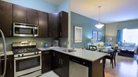 This single-floor residence offers a thoughtfully designed open-concept layout that seamlessly connects the kitchen and dining areas, creating a cohesive and inviting living environment. The kitchen is a standout feature, showcasing rich dark espresso cabinetry that provides generous storage and a striking visual contrast against the cool blue-grey accent walls. Granite countertops extend across the workspace and wrap around a peninsula island, which includes an undermount sink with a contemporary arc faucet and integrated dishwasher. Stainless steel appliances, including a gas range and built-in microwave, add a modern, professional touch. A stylish pendant light hangs above the peninsula, enhancing both function and ambiance. The adjacent dining area flows naturally from the kitchen, illuminated by a matching pendant fixture and adorned with framed wall art, lending the space a curated, finished feel. Light-toned flooring runs throughout, brightening the interior and complementing the warm cabinetry tones. The overall aesthetic balances contemporary design with everyday practicality, making this a comfortable and visually appealing space suited for both daily living and entertaining.