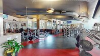 Located at 3311 Woodhill Ln in Fayetteville, NC, this single-story fitness facility combines industrial-modern architecture with a comprehensive range of workout amenities. The space is defined by its high ceilings with exposed ductwork, pendant lighting, and a dark ceiling that creates a dynamic, energetic atmosphere throughout.

The main workout floor is divided into distinct zones, featuring durable rubber flooring with red and gray sections that delineate areas for free weights, strength machines, and cardio equipment. Exposed brick columns punctuate the space, adding a raw, urban character that contrasts with the clean, modern finishes elsewhere.

The free weight area is generously equipped with multiple dumbbell racks, adjustable benches, and weight machines, all reflected in full-length wall mirrors that enhance the sense of openness. The cardio section, positioned along a wall of large floor-to-ceiling windows, houses treadmills, ellipticals, stationary bikes, and stair climbers, bathed in abundant natural light with views of the surrounding landscape.

A distinctive reception and transition area features a sculptural staircase with bold yellow accents, a raised planter filled with lush greenery, and decorative elements that add a welcoming, vibrant focal point. Mounted televisions and motivational artwork are positioned throughout, completing this well-appointed and thoughtfully designed fitness center.