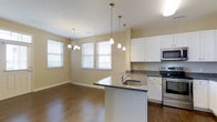This single-floor residence is thoughtfully designed to maximize both function and comfort. A welcoming hallway with dark hardwood flooring and clean white trim leads the way into the main living areas, framed by an elegant arched entryway that adds a refined architectural touch. Paneled doors and neutral warm-toned walls contribute to a cohesive, polished aesthetic throughout.

The heart of the home is the open-concept kitchen and dining area, where rich dark hardwood floors continue seamlessly underfoot. The kitchen is outfitted with crisp white cabinetry, dark granite countertops, and a full suite of stainless steel appliances, including a range and microwave. A central island with an integrated sink serves as both a practical workspace and a natural gathering point. Pendant lighting hangs above the island, while a classic chandelier illuminates the adjacent dining area, which benefits from generous natural light streaming through multiple large windows dressed with horizontal blinds. The open layout between the kitchen and dining space creates an airy, connected feel that is well-suited for both everyday living and entertaining.