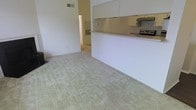 This single-floor space features an open-concept layout that integrates the living area and kitchen into one cohesive environment. The living area is finished with light-toned wall-to-wall carpeting, lending a soft, neutral aesthetic throughout. A half-wall partition separates the living space from the kitchen, topped with a light-colored countertop that doubles as a convenient breakfast bar or pass-through surface. The kitchen is equipped with white cabinetry mounted above, providing ample storage, along with a range and an overhead exhaust hood. The walls are painted in a clean, bright white, contributing to an airy and open feel. A doorway visible in the background connects to an additional room, suggesting practical flow between spaces. Ventilation is supported by a ceiling vent, and electrical outlets are strategically placed along the walls. The overall design is straightforward and functional, with a neutral color palette that offers flexibility for a variety of furnishing styles. The layout maximizes usable floor space while maintaining clear sightlines across the living and kitchen areas.