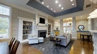 Located at 10 Primrose in Haverhill, Massachusetts, this single-floor space exudes a refined and welcoming ambiance through its thoughtful architectural design and upscale finishes. The centerpiece of the main lounge area is a classic fireplace framed by a crisp white mantel and flanked by built-in shelving units, offering both elegance and functional display space. Recessed lighting illuminates the room from above, complementing the distinctive tray ceiling detail accented in a sophisticated slate blue tone that adds depth and visual interest. Decorative columns frame an archway that connects to an adjacent area, where a chandelier and large windows invite natural light deep into the space. Rich hardwood flooring anchors the room, transitioning seamlessly to a patterned area rug that defines the seating area. Crown molding, wainscoting-style wall panels, and neutral tones throughout contribute to a polished, classic aesthetic. The layout flows generously, creating an open yet intimate atmosphere ideal for gathering and relaxation. Overall, the space balances traditional architectural charm with modern comfort.