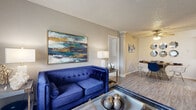 This single-floor residence is thoughtfully designed with a cohesive navy, teal, and gold aesthetic carried throughout its interior spaces. The living area serves as the heart of the home, featuring wood-look flooring, neutral walls, and a textured ceiling. A tufted navy velvet sofa anchors the space, complemented by a glass coffee table and bold abstract wall art with teal and gold tones. The living area flows openly into a dedicated dining space, maintaining a connected, sociable layout.

The galley-style kitchen is efficiently organized, offering dark cabinetry with granite-look countertops, a stainless steel refrigerator, electric range, and dishwasher. Wood-look flooring continues through the kitchen, adding visual continuity.

Two bedrooms provide comfortable personal retreats. The primary bedroom features carpet flooring, a large window with teal drape panels, and a spacious walk-in closet with built-in shelving. The secondary bedroom also features carpeted floors and a bright window. A full bathroom includes a tub-shower combination, dark wood vanity with granite-style countertop, and wood-look tile flooring. A dedicated utility area with a water heater rounds out the functional aspects of the space.