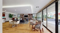 This thoughtfully designed single-floor amenity space offers a seamless blend of comfort, function, and contemporary style. The main lounge area is anchored by a striking floor-to-ceiling built-in bookcase flanking a stone-framed fireplace and wall-mounted television, creating a warm and inviting focal point. Warm-toned hardwood flooring flows throughout, complementing the clean white ceilings with recessed lighting and a subtle tray ceiling detail that adds architectural depth. The lounge is furnished with a versatile mix of seating arrangements — from casual swivel chairs and a patterned area rug to dining-height tables and upholstered chairs — accommodating both individual relaxation and group gatherings. Floor-to-ceiling sliding glass doors line an entire wall, flooding the interior with abundant natural light and providing direct access to an outdoor patio area with additional seating. A dedicated bar and counter area features a sleek wall-mounted ledge with stylish bar stools and curated artwork above. The kitchenette is a standout feature, boasting rich walnut cabinetry, a vibrant turquoise tile backsplash, white quartz countertops with a sculptural curved island, open shelving stocked with glassware, a coffee station, microwave, and stainless steel appliances — making this space fully equipped for entertaining and everyday use.