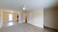 This two-floor residence presents a clean, practical layout designed for comfortable everyday living. The image captures what appears to be a bedroom or main living area, finished with wall-to-wall neutral-toned carpeting that adds warmth underfoot. A ceiling fan with a built-in light fixture is centrally mounted on the textured ceiling, providing both airflow and ambient lighting to the space.

Two white interior doors are positioned along the far wall, one of which opens to reveal a bathroom with warm lighting, while the other appears to lead to a closet or additional storage space. An open doorway on the left side of the room offers a glimpse into an adjacent area, where cabinetry and what appears to be kitchen or utility storage can be seen in the background.

The walls are finished in a soft, neutral cream tone that complements the light carpeting and white trim throughout. A staircase is partially visible along the right edge of the frame, confirming the multi-level nature of the property. The overall ambiance is understated and move-in ready, with a straightforward design that prioritizes function and ease of use across both floors.