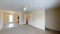 This two-floor residence presents a clean, practical layout designed for comfortable everyday living. The image captures what appears to be a bedroom or main living area, finished with wall-to-wall neutral-toned carpeting that adds warmth underfoot. A ceiling fan with a built-in light fixture is centrally mounted on the textured ceiling, providing both airflow and ambient lighting to the space.

Two white interior doors are positioned along the far wall, one of which opens to reveal a bathroom with warm lighting, while the other appears to lead to a closet or additional storage space. An open doorway on the left side of the room offers a glimpse into an adjacent area, where cabinetry and what appears to be kitchen or utility storage can be seen in the background.

The walls are finished in a soft, neutral cream tone that complements the light carpeting and white trim throughout. A staircase is partially visible along the right edge of the frame, confirming the multi-level nature of the property. The overall ambiance is understated and move-in ready, with a straightforward design that prioritizes function and ease of use across both floors.