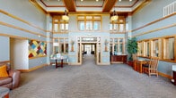 This single-floor facility presents a thoughtfully designed interior that balances professional functionality with welcoming comfort. The grand lobby serves as the centerpiece of the space, boasting soaring double-height ceilings, warm wood-trimmed transom windows, and soft blue-gray walls that create an airy, upscale atmosphere. Pendant lighting and decorative accents, including colorful geometric artwork, add character to the entrance area.

Adjacent seating areas feature plush leather chairs and sofas arranged beneath an elegant multi-arm chandelier, with floor-to-ceiling windows flooding the space with natural light and offering views to the outdoors. Warm wood trim and crown molding are consistent design elements throughout, lending a cohesive craftsman aesthetic to the entire facility.

The fully equipped kitchen showcases rich cherry wood cabinetry, dark granite countertops, stainless steel appliances, and a large central island with bar seating — ideal for communal gatherings or catered events. A dedicated business center provides multiple computer workstations along a continuous desk, framed artwork, and vibrant olive and blue accent walls. Wide carpeted corridors with high ceilings connect the various functional rooms and private spaces, all finished with the same warm wood millwork and soft neutral tones that define this inviting and versatile facility.