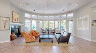 Located at 1500 Seagull Dr in Palm Harbor, Florida, this well-appointed single-floor community space offers a thoughtfully designed interior that balances function and comfort. Light wood-look flooring flows seamlessly throughout, complementing the soft neutral gray walls and crisp white trim found in every area.

The expansive living room serves as the heart of the space, featuring soaring ceilings with transom windows that flood the interior with natural light. A striking dark marble fireplace with a white mantel anchors one wall, while a sweeping curved window wall offers direct views of the outdoor amenity area. Elegant decorative columns frame transitions between zones, adding architectural character.

The dining area is an intimate, naturally lit nook defined by an arched window and additional clerestory windows above, creating a bright and welcoming atmosphere for gatherings. Adjacent to it, a versatile secondary dining and seating area is surrounded by large windows on multiple sides, inviting views of lush greenery.

A dedicated workspace area accommodates workstations and office equipment, lined with a row of high clerestory windows that provide even, glare-free light. A curved sunroom-style sitting area, enclosed by floor-to-ceiling windows, rounds out the space with panoramic outdoor views and an airy, open ambiance throughout.