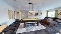 This single-floor game room offers a well-appointed social space centered around a substantial dark-wood pool table with a striking yellow felt surface. Suspended above it, an industrial-style pendant light fixture with exposed Edison bulbs provides focused illumination while adding a modern-rustic design touch. The room's walls are clad in light-toned horizontal shiplap paneling, lending a clean, contemporary aesthetic throughout the space.

Along one wall, a flat-screen television is mounted above a series of rustic floating wooden shelves displaying small decorative accents, creating an inviting media area. Adjacent to this, a brown leather sofa offers comfortable seating for loungers and spectators alike. The flooring features a patterned carpet tile in muted tones, with a large area rug anchoring the pool table at the room's center.

Recessed ceiling lighting complements the pendant fixture, ensuring the space is well-lit throughout. A cue rack is mounted near the entrance, keeping equipment accessible and organized. The overall ambiance balances casual comfort with recreational function, making it a versatile gathering space suited for social interaction and leisure activities.