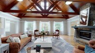 Located at 809 E Main St in Lexington, SC, this single-floor community clubhouse is a beautifully designed gathering space that blends rustic architectural character with comfortable, contemporary furnishings. The centerpiece of the main lounge is a soaring vaulted ceiling clad in warm wood planking and supported by richly stained exposed timber trusses, creating a dramatic and welcoming atmosphere. Clerestory windows along the upper walls flood the space with abundant natural light, while large double-hung windows dressed in sheer white curtains frame lush outdoor views along the perimeter. A floor-to-ceiling stacked stone fireplace anchors one wall of the main seating area, which features leather sofas, tufted armchairs, and an upholstered ottoman arranged over patterned area rugs atop hardwood floors. Adjacent to the main lounge, a secondary lounge area offers vaulted ceilings with ceiling fans, plush curved seating, and a wall-mounted television, providing a more casual retreat. A bright café-style nook with a high-top table, bar seating, and pendant lighting offers a quiet spot for small gatherings. The entry foyer, finished with hardwood floors and a built-in credenza, provides a welcoming transition into the space. Throughout, neutral wall tones and consistent trim work tie the various areas together cohesively.