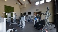 Located at 740 Bridgeport Ave in Shelton, Connecticut, this fitness center offers a spacious, single-floor layout designed to accommodate a full range of exercise needs. The space is defined by its striking vaulted ceiling with exposed dark wood beams, which adds architectural character and contributes to an open, airy atmosphere. Clerestory windows positioned high along the upper walls allow natural light to filter in, brightening the interior throughout the day. Pendant lighting and recessed fixtures provide additional illumination.

The fitness floor is finished with durable dark rubber flooring, providing a functional surface suited for both cardio and strength training activities. The equipment selection includes Life Fitness cardio machines such as ellipticals and stationary bikes, as well as multi-station strength machines and free weights. Wall-mounted flat-screen televisions are positioned for easy viewing during workouts.

A dedicated area toward the rear of the space, visible through a doorway, appears to offer additional workout options, including stability balls and supplementary equipment. A decorative living green wall accent adds a natural, refreshing element to the overall design, enhancing the ambiance of the space.