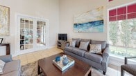 This single-floor space is defined by its striking architectural height, with soaring ceilings that create an expansive, airy ambiance throughout. The open-concept layout seamlessly connects the main living, dining, and kitchen areas, offering a cohesive and functional flow.

The living area is generously proportioned, featuring large multi-pane windows dressed with bold crimson roller shades that allow ample natural light while adding a vibrant design accent. Transom windows positioned high on the walls further amplify the sense of volume and brightness. French doors with glass panels open to an adjacent room, enhancing the openness of the space.

The dining area accommodates a substantial dark wood table surrounded by upholstered white nail-head chairs, anchored by a large abstract landscape artwork on the wall above. Light tile flooring runs consistently throughout, contributing to the clean, cohesive aesthetic.

The kitchen is well-equipped with warm wood cabinetry, dark granite countertops, a double sink, and stainless steel appliances including a side-by-side refrigerator. A separate room accessed through elegant double French doors features glass panes, offering visibility into what appears to be a dedicated office or study space. Overall, the space balances practicality with refined, contemporary styling.