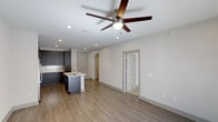 This single-floor residence in Plano offers a thoughtfully designed open-concept layout that seamlessly connects the kitchen and living areas. The kitchen is a standout feature, showcasing dark cabinetry with a contemporary finish, a classic subway tile backsplash, a stainless steel sink set within a functional center island, and modern appliances. The warm hardwood-style flooring flows continuously throughout the space, adding a sense of cohesion and elegance. Recessed lighting is strategically placed across the ceiling, providing clean, even illumination, while a ceiling fan with a brushed nickel finish serves as both a practical and stylistic centerpiece above the main living area. The neutral, light-toned walls create a bright and welcoming ambiance, offering a versatile backdrop for a variety of interior styles. Doorways lead to additional rooms, including what appears to be a walk-in closet and a bathroom, suggesting a well-organized and functional floor plan. The overall design strikes a balance between modern aesthetics and everyday practicality, making this Plano residence a comfortable and stylish living environment.