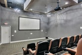 Located in Murrieta, CA, this dedicated screening room delivers an authentic cinema experience within a compact, well-appointed space. The room is designed with tiered, stadium-style seating arranged in multiple rows, each occupied by plush tan leather chairs with built-in armrests and cup holders — providing both comfort and functionality for viewers. The stepped floor layout ensures clear sightlines from every seat to the large projection screen mounted on the front wall. A ceiling-mounted projector is centrally positioned to deliver a sharp, full-sized image onto the screen. The interior features neutral gray walls with subtle textured finishes, accented by a patterned ceiling treatment that adds visual depth and aids in acoustic performance. Elegant chandelier-style pendant lights are suspended from a coffered ceiling detail, complementing recessed lighting throughout. The flooring is finished with a striped, commercial-grade carpet that runs the length of the room, reinforcing the theater aesthetic. LED step lighting along the tiered aisles ensures safe navigation in low-light conditions. The space connects to a hallway corridor, providing organized entry and exit flow. Overall, the room offers a polished, immersive screening environment well-suited for private viewings or community entertainment.