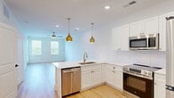 Located at 7015 Ruth Ave in Wilmington, NC, this thoughtfully designed one-bedroom, one-bathroom home offers a clean, modern aesthetic throughout its well-utilized single floor. The open-concept kitchen and living area serves as the heart of the home, featuring crisp white shaker cabinets adorned with gold hardware, a classic white subway tile backsplash, and a full suite of stainless steel appliances including a refrigerator, range, built-in microwave, and dishwasher. Two eye-catching gold pendant lights hang above the kitchen peninsula, adding a warm, contemporary touch. Light-toned hardwood flooring flows through the main living areas, complementing the bright white walls and creating an airy, inviting atmosphere. The living room benefits from natural light through windows with blinds and is equipped with a ceiling fan for year-round comfort. A glass-panel exterior door provides additional natural light and outdoor access. The generously sized bedroom features soft carpeting, a ceiling fan, and a window with outdoor views, creating a calm and restful retreat. The full bathroom is finished with hardwood-style flooring, white cabinetry with a vanity mirror, and a full bathtub. A hallway connects the spaces and includes closet storage, completing this efficient and stylish home.