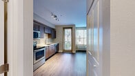 This well-appointed single-floor residence showcases a thoughtfully designed open layout that seamlessly connects the kitchen and living areas. The kitchen is a functional highlight, featuring a full suite of stainless steel appliances—including a refrigerator, electric range, built-in microwave, and dishwasher—paired with warm wood-toned cabinetry, light countertops, and a contemporary sink with a modern faucet. Track lighting overhead ensures excellent task illumination throughout the cooking space.

The living area benefits from generous natural light streaming through large windows fitted with horizontal blinds, as well as direct access to a private balcony through a floor-to-ceiling glass door. A wall-mounted HVAC unit provides year-round climate control. Consistent hardwood-style flooring flows throughout the space, lending a cohesive and polished aesthetic.

Additional rooms include a well-sized bedroom with neutral walls, hardwood-style flooring, and ample natural light, providing a calm and comfortable retreat. The bathroom is clean and practical, finished with light wood-look flooring and crisp white fixtures. Overall, the space offers a modern, functional design with quality finishes and a bright, welcoming ambiance throughout.