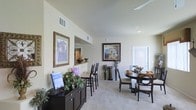 This single-story space showcases an open-plan layout that seamlessly integrates multiple living and dining areas. The interior is characterized by elevated ceilings that enhance the sense of volume throughout. Neutral, light-toned walls create a bright and airy ambiance, complemented by wall-to-wall carpeting that adds warmth underfoot.

A formal dining area anchors one side of the space, furnished with a round dark-wood table and upholstered chairs. Adjacent to it, a bar-height counter with tall chairs provides a casual seating alternative, overlooking a pass-through opening that connects to a neighboring room — likely a kitchen. This architectural detail adds both functionality and visual interest to the layout.

Decorative framed artwork adorns the walls, contributing an elegant, curated aesthetic. A dark-wood sideboard positioned along one wall offers additional storage and display space, styled with decorative plants and accent pieces. Recessed lighting and ceiling-mounted vents are integrated throughout, ensuring comfort and even illumination. A door with sidelight panels at the far end suggests access to an exterior area, allowing natural light to filter into the space.