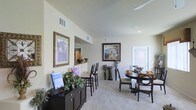 This single-story space showcases an open-plan layout that seamlessly integrates multiple living and dining areas. The interior is characterized by elevated ceilings that enhance the sense of volume throughout. Neutral, light-toned walls create a bright and airy ambiance, complemented by wall-to-wall carpeting that adds warmth underfoot.

A formal dining area anchors one side of the space, furnished with a round dark-wood table and upholstered chairs. Adjacent to it, a bar-height counter with tall chairs provides a casual seating alternative, overlooking a pass-through opening that connects to a neighboring room — likely a kitchen. This architectural detail adds both functionality and visual interest to the layout.

Decorative framed artwork adorns the walls, contributing an elegant, curated aesthetic. A dark-wood sideboard positioned along one wall offers additional storage and display space, styled with decorative plants and accent pieces. Recessed lighting and ceiling-mounted vents are integrated throughout, ensuring comfort and even illumination. A door with sidelight panels at the far end suggests access to an exterior area, allowing natural light to filter into the space.
