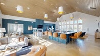 Located at 1001 NW Cernan Dr in Huntsville, Alabama, this expansive single-floor community space offers a generously sized, open-concept living room designed for both relaxation and social interaction. The vaulted ceiling, clad in warm natural wood planking, rises dramatically overhead and is complemented by stylish pendant lights with gold-accented shades, creating an inviting and airy atmosphere throughout.

The interior is defined by bold teal-blue paneled walls that anchor the space with a distinctive coastal-modern aesthetic. A central fireplace with a chevron-tiled surround and wall-mounted television serves as the focal point of the main lounge area, which features plush seating, a sectional sofa, and layered rugs over polished concrete floors. An adjacent bar counter, wrapped in matching teal paneling and lined with cognac leather barstools, provides a communal gathering spot.

Multiple activity zones are thoughtfully integrated within the open floor plan, including a ping-pong table, a billiards table, and a workspace alcove. Floor-to-ceiling gridded windows along one wall flood the interior with abundant natural light and frame outdoor views of the surrounding neighborhood. The combination of warm wood tones, rich teal accents, and bright white walls gives the space a cohesive, modern, and welcoming character.
