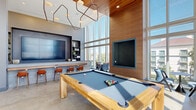 This contemporary amenity lounge is designed to impress, offering a dynamic and versatile space for recreation and relaxation. The room is anchored by dramatic double-height ceilings clad in warm wood paneling, which contrasts beautifully with the polished concrete floors below. Floor-to-ceiling windows wrap the space in abundant natural light while framing views of the surrounding urban landscape.

At the heart of the room sits a solid wood billiards table with a slate-blue felt surface, positioned beneath a striking sculptural chandelier. A wall-mounted flat-screen TV is recessed into the wood-paneled accent wall, complemented by a second large display integrated into the opposite wall. A built-in bar counter with bar-height seating and open shelving provides a convenient refreshment area.

The lounge zone features a generously sized sectional sofa in soft blue-grey upholstery arranged around low round coffee tables, ideal for social gatherings. A sleek kitchenette with stone countertops, an undermount sink, warm wood cabinetry, and a built-in microwave serves the space efficiently. Glass doors open directly to an outdoor terrace equipped with a gas grill, extending the entertaining space outdoors. Colorful abstract artwork adds a curated, gallery-like touch throughout.
