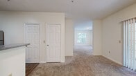 This single-floor residence offers a thoughtfully arranged layout comprising one bedroom, one bathroom, a living room, a hallway, and a dedicated closet. Soft, neutral tones and plush wall-to-wall carpeting flow throughout the main living areas, creating a warm and cohesive ambiance. The open living room is generously proportioned and features a sliding glass door fitted with vertical blinds, providing direct access to an outdoor balcony and inviting an abundance of natural light along with pleasant greenery views. The kitchen is positioned adjacent to the living area and features a breakfast bar-style pass-through counter, light wood cabinetry, a stainless steel refrigerator, and wood-look flooring that adds a clean contrast to the carpeted spaces. The bedroom is a comfortable, well-lit retreat with large windows dressed in horizontal blinds that frame lush outdoor tree views. It connects directly to a walk-in closet, offering practical storage. The bathroom is finished with wood-look flooring, light wood cabinetry, a wide vanity with a speckled granite-look countertop, an undermount sink, and a large mirror. Throughout the space, clean white walls, textured ceilings, and classic paneled doors contribute to a simple, functional, and move-in-ready aesthetic.