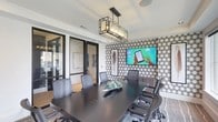 This single-floor conference room is a well-appointed meeting space designed with a contemporary aesthetic and functional layout. The room's most striking feature is its bold black-and-white hexagonal patterned accent wall, which serves as a dynamic backdrop to a large wall-mounted display screen ideal for presentations and video conferencing. Flanking the screen, framed feather artwork adds an artistic touch that softens the geometric pattern.

At the center of the room sits a substantial dark conference table surrounded by ergonomic mesh-back chairs, comfortably accommodating groups for collaborative work sessions. Built-in power access points are integrated directly into the table surface for added convenience.

Overhead, an industrial-style rectangular pendant light fixture with exposed bulbs anchors the space, providing both task lighting and a design focal point. Large windows along one wall flood the room with natural light while offering views of the exterior landscaping and surrounding streetscape. The patterned area rug beneath the table adds texture and warmth, complementing the neutral walls and light wood flooring visible near the glass-paneled entry doors, which connect seamlessly to adjacent spaces.