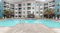 Located at 1400 Encore Pl in Sanford, Florida, this vibrant outdoor amenity courtyard is the centerpiece of a modern multi-story residential community. The space is anchored by a sparkling resort-style swimming pool complete with a dramatic waterfall feature, surrounded by expansive stone-paved pool decking. Rows of sleek white lounge chairs line the sun deck, offering ample space for relaxation alongside lush tropical landscaping featuring towering palm trees, ferns, and dense tropical plantings that enhance the Florida ambiance.

A covered lounge area framed by striking natural stone columns provides a shaded retreat, furnished with wicker seating and teal accent cushions, creating a comfortable outdoor living space. Teal market umbrellas add pops of color throughout the courtyard, complementing the building's bold blue and gray exterior cladding. The multi-story residential building wraps around the courtyard on multiple sides, with balconies overlooking the amenity space, offering residents direct views of the pool and greenery below.

Paved walkways connect the various zones of the courtyard seamlessly, while carefully curated planting beds and mature palms lend a resort-like atmosphere. The overall design blends contemporary architecture with tropical landscaping, creating an inviting and functional outdoor community hub.