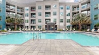 Located at 1400 Encore Pl in Sanford, Florida, this vibrant outdoor amenity courtyard is the centerpiece of a modern multi-story residential community. The space is anchored by a sparkling resort-style swimming pool complete with a dramatic waterfall feature, surrounded by expansive stone-paved pool decking. Rows of sleek white lounge chairs line the sun deck, offering ample space for relaxation alongside lush tropical landscaping featuring towering palm trees, ferns, and dense tropical plantings that enhance the Florida ambiance.

A covered lounge area framed by striking natural stone columns provides a shaded retreat, furnished with wicker seating and teal accent cushions, creating a comfortable outdoor living space. Teal market umbrellas add pops of color throughout the courtyard, complementing the building's bold blue and gray exterior cladding. The multi-story residential building wraps around the courtyard on multiple sides, with balconies overlooking the amenity space, offering residents direct views of the pool and greenery below.

Paved walkways connect the various zones of the courtyard seamlessly, while carefully curated planting beds and mature palms lend a resort-like atmosphere. The overall design blends contemporary architecture with tropical landscaping, creating an inviting and functional outdoor community hub.