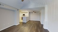 This single-story space showcases a thoughtfully designed open-concept layout that seamlessly connects the living area and kitchen. Rich, dark hardwood-style flooring runs throughout, providing a warm contrast against the crisp white walls and bright, neutral color palette. The living area is generously proportioned, offering flexible furniture arrangement options, and is anchored by a ceiling fan with a built-in light fixture that enhances both comfort and illumination. Vaulted ceiling detailing adds an architectural dimension to the space, contributing to an airy, open feel. The kitchen features a peninsula with countertop seating, white cabinetry, stainless steel appliances including a microwave, and recessed lighting overhead. The peninsula creates a natural division between the kitchen and living areas while maintaining the open flow of the floor plan. A sliding door is visible along one wall, potentially leading to additional storage or a closet. The overall ambiance is clean, modern, and move-in ready, with a neutral design palette that allows for easy personalization.