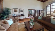 This sophisticated single-floor space showcases exceptional architectural craftsmanship throughout. Soaring vaulted ceilings with decorative wooden beams and arched detailing create a grand sense of openness, while warm hardwood floors flow seamlessly from room to room, complemented by rich dark wood trim and baseboards that frame every doorway and window.

The main living area serves as the heart of the space, anchored by a classic fireplace with an ornate wood mantel and framed artwork above. Comfortable tufted and upholstered seating arrangements are centered around wooden coffee tables, lending a welcoming and refined ambiance. A console table with decorative accents and framed landscape art adds to the curated aesthetic.

An adjacent seating area benefits from dramatic floor-to-ceiling windows that flood the space with natural light and offer views of an outdoor stone-accented terrace. Dark-framed French doors and paneled solid wood doors enhance the traditional character throughout.

Arched colonnades with decorative columns define transitions between areas, adding architectural depth and visual interest. Recessed lighting and pendant fixtures provide warm illumination that highlights the space's refined details. The overall atmosphere is one of timeless elegance, combining classic design elements with an inviting, well-appointed interior suited for both relaxed gatherings and formal occasions.