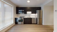 Located at 1500 Hancock St in Quincy, Massachusetts, this modern single-floor residence offers a thoughtfully designed layout with contemporary finishes throughout. The open-concept living and dining area is finished with light wood-look flooring and crisp white walls, creating a bright and inviting atmosphere. Multiple large windows flood the space with natural light and provide views of the surrounding area, while stylish cylindrical pendant lights add a contemporary touch to the ceiling.

The kitchen is a standout feature, showcasing sleek dark cabinetry that contrasts sharply with white countertops and a classic subway tile backsplash. A full suite of stainless steel appliances — including a range, microwave, dishwasher, and refrigerator — provides a highly functional cooking environment that flows seamlessly into the main living space.

The unit includes two bathrooms, each finished with wood-look tile flooring and dark vanity cabinetry with white countertops. One bathroom features a full soaking tub with shower combination, while the second offers a dedicated walk-in shower stall, providing versatility for residents. The floor plan also includes bedroom space with carpeted flooring, offering a comfortable contrast to the hard surfaces found elsewhere in the unit. Overall, the space presents a clean, modern aesthetic well-suited for everyday living.