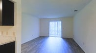 This single-story space presents a clean, minimalist interior designed to maximize functionality and comfort. The main living area is finished with smooth, wood-style flooring in a cool gray-brown tone, lending the space a modern and polished aesthetic. The floors reflect natural light effectively, contributing to an open and airy atmosphere throughout the room.

The walls are finished in a neutral white, providing a versatile backdrop suitable for a variety of interior design styles. A standout feature of this space is the full-width sliding glass door positioned along one wall, fitted with vertical blinds that allow for adjustable light control. This door serves as the primary source of natural light, brightening the interior and offering a visual connection to the outdoors.

The ceiling is smooth and standard in height, maintaining a consistent and uncluttered overhead appearance. The overall layout of the main room appears open and unobstructed, offering flexible possibilities for furniture arrangement. The combination of neutral tones, natural light, and wood-style flooring creates a simple yet functional living environment.