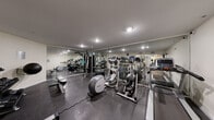 This fitness center is designed to offer a complete workout experience within a single, efficiently organized space. The room features durable rubberized flooring throughout, providing both safety and comfort during exercise. Floor-to-ceiling mirrors line the walls, visually expanding the space while allowing users to monitor their form during workouts.

The cardio section is well-stocked with multiple treadmills, elliptical trainers, and a stationary bike, offering a variety of aerobic exercise options. A multi-station cable machine anchors the strength training area, complemented by a dedicated free weights section that includes a dumbbell rack and a medicine ball. Flat weight benches provide additional versatility for strength routines.

Recessed and flush-mounted ceiling lights ensure the space is evenly and brightly illuminated throughout. Informational signage is posted throughout the room, and a wall clock helps users track their workout sessions. The facility includes a private restroom with a vanity, mirror, and all essential amenities for post-workout convenience.

The overall layout is clean and functional, with equipment arranged to maximize usable floor space and allow for comfortable movement between stations. The neutral color palette of white walls and gray tones lends the space a modern, professional atmosphere.