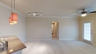This single-story space presents a well-maintained interior defined by a neutral, warm color palette throughout. The main living area features plush wall-to-wall carpeting in a light beige tone, contributing to a quiet and comfortable atmosphere. Walls are finished in a soft, warm white that complements the clean white baseboards and crown molding, adding a subtle architectural refinement to the room.

Overhead, a track lighting system with adjustable spotlights provides flexible, directed illumination, making it easy to highlight different areas of the room as needed. The ceiling is smooth and consistent, with crown molding detail that runs along the perimeter, lending a polished finish to the space.

A standout architectural feature is the rounded arched entryway that connects this main room to an adjacent hallway or secondary space. This graceful arch adds visual interest and a classic design touch, breaking up the otherwise straightforward layout. The adjoining corridor, visible through the arch, continues the same neutral tones and carpeting, ensuring a cohesive aesthetic throughout the space.

Overall, the space offers a clean, move-in ready environment with classic detailing and a calm, understated ambiance.