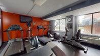 Located at 600 Asylum Ave in Hartford, Connecticut, this dedicated fitness center is designed to support a well-rounded workout experience. The space is finished with durable rubber flooring throughout and features a bold color scheme, with a striking orange accent wall serving as the visual centerpiece. Motivational phrases such as "Challenge" and "Change" are displayed prominently on the wall, reinforcing an energizing atmosphere.

The cardio section is well-equipped, offering multiple treadmills, elliptical trainers, and stationary bikes from brands including TRUE and Matrix, all arranged to maximize use of the floor space. A wall-mounted flat-screen television is positioned above the orange accent wall, providing entertainment during workouts. A multi-function cable strength training machine and an adjustable workout bench occupy a dedicated strength training area, complemented by a large wall mirror that enhances the sense of space.

Natural light flows into the room through several windows along the gray perimeter walls, offering views of the surrounding outdoor greenery. A wall clock and posted fitness center rules keep users informed and on schedule. The space also includes a private restroom accessible via a door at the rear. The overall design is functional and motivating, making it a practical facility for daily fitness routines.