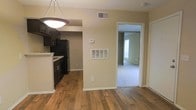 This single-floor residence offers a well-organized layout that balances functionality and comfort. The main living area is anchored by warm, rich hardwood flooring that extends throughout the open-concept space, lending a classic and inviting character to the interior. A pendant light fixture adds a focused design element overhead, complementing the neutral, beige-toned walls that keep the atmosphere bright and understated.

The kitchen is positioned adjacent to the living area and features dark cabinetry with ample storage, a black refrigerator, and a light-colored countertop that provides a clean contrast to the darker finishes. A raised counter divides the kitchen from the living space, offering a natural transition between the two areas.

Climate control is well-considered, with both a ceiling vent and a wall-mounted HVAC return visible in the main area, ensuring consistent airflow throughout the space. A wall-mounted thermostat allows for convenient temperature management.

Through an open doorway, the bedroom is visible and transitions to plush carpeting, offering a softer, quieter environment for rest. The room features a closet and a window that allows natural light to filter in. White trim and baseboards throughout the space provide a crisp, finished look that ties the overall aesthetic together cohesively.