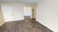 This single-floor space presents a straightforward, functional layout characterized by clean lines and neutral finishes throughout. The flooring consists of wood-look planks in a warm medium-brown tone, running consistently across the open area and continuing into an adjoining room visible through a wide doorway. The walls are finished in a soft, warm off-white, complementing the light-toned baseboards that frame the perimeter of each room. The ceiling features a classic textured popcorn finish, adding subtle depth overhead.

A notable architectural element is the tall, vertical floor-to-ceiling HVAC register unit positioned near the doorway threshold, suggesting a wall-mounted heating and cooling system. The wide, open passage between the two visible rooms creates a sense of connected, flowing space, making the layout feel accessible and easy to navigate. Electrical outlets and switch plates are positioned at standard intervals along the walls, indicating the space is move-in ready. The overall ambiance is clean, bright, and neutral, providing a blank canvas suitable for a variety of uses and furnishing styles.